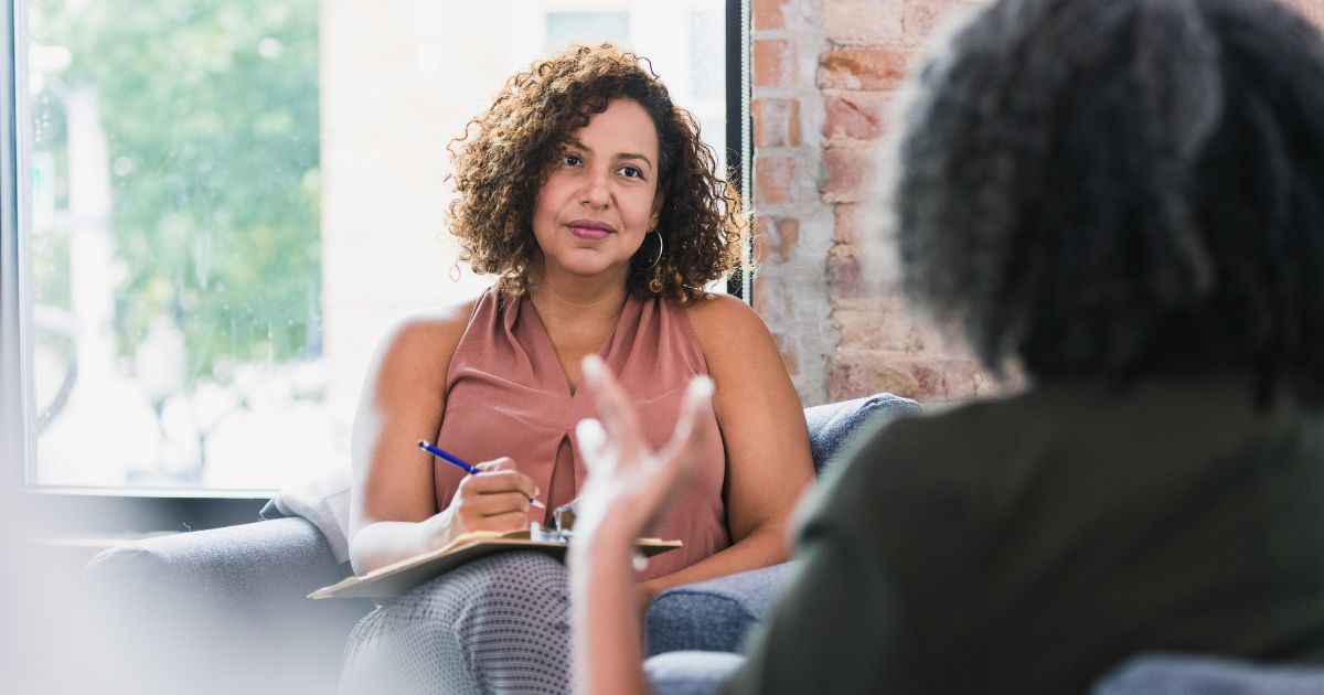 A female worker with curly hair is listening to her client talking and taking notes as part of her mental health career duties.