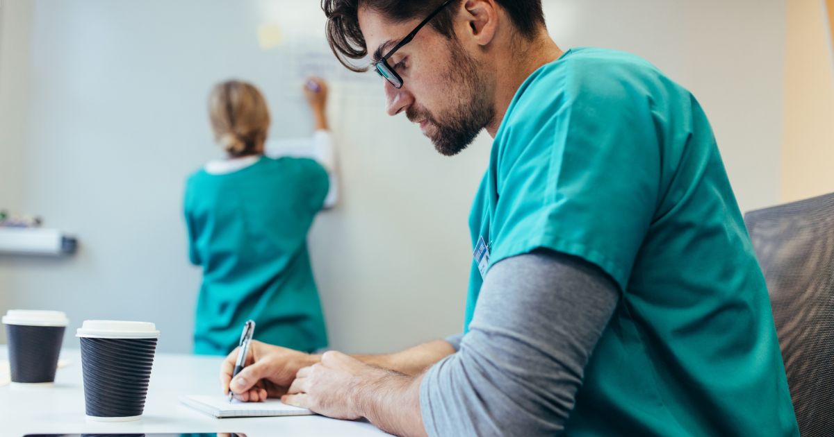Healthcare worker sitting at his desk writing notes. He could be working in the following health jobs: health assistant or health administrator.